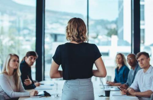 A woman in a black shirt and grey pants leads a business meeting. Five people sit at the table listening to her talk.
