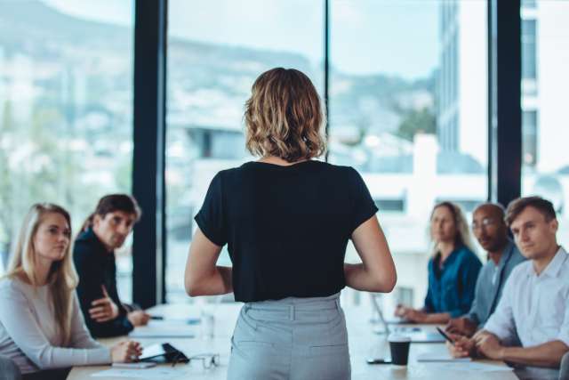 A woman in a black shirt and grey pants leads a business meeting. Five people sit at the table listening to her talk.