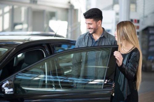 A young man and woman stand near a car with the driver-side door open, looking in and smiling in a dealership showroom.