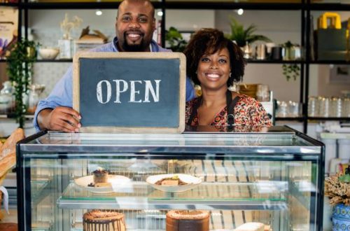 Two entrepreneurs in their newly opened dessert shop, standing behind a display case full of cakes and pastries.