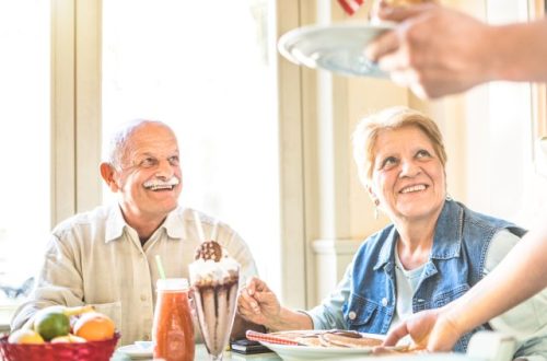 A senior man and woman sit at a table filled with a milkshake and breakfast food. They look sideways at their server.