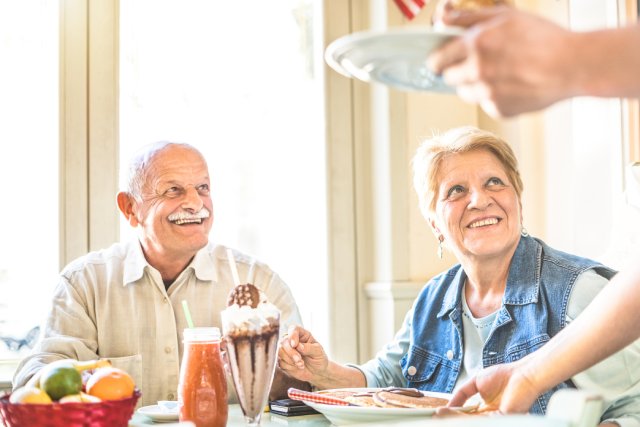 A senior man and woman sit at a table filled with a milkshake and breakfast food. They look sideways at their server.