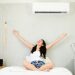 A happy young woman with long black hair stretches out with joy on her bed underneath her mini split unit.