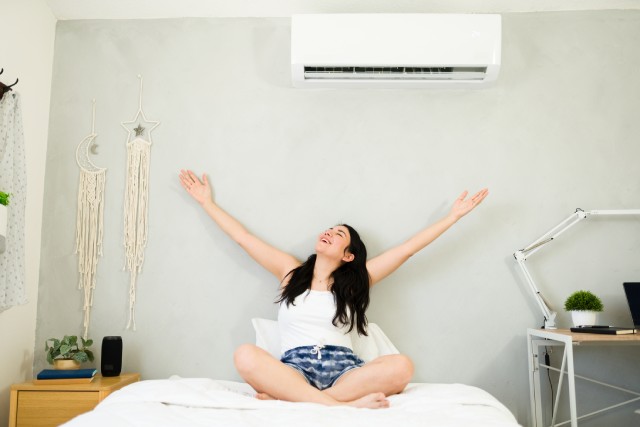 A happy young woman with long black hair stretches out with joy on her bed underneath her mini split unit.
