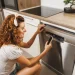 A young woman crouching in front of her smart dishwasher. She has one hand on the appliance door and a phone in the other.