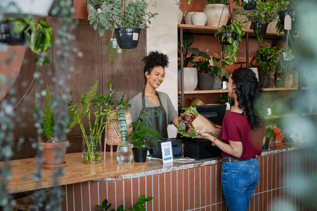 A young woman stands behind a counter in a black apron and gray shirt and hands a woman a floral bouquet.