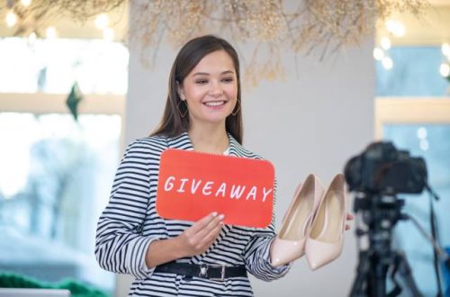 A smiling brunette woman holds a pair of shoes and a sign with the word "Giveaway" as she faces a camera on a tripod.