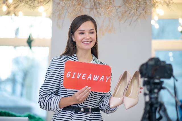 A smiling brunette woman holds a pair of shoes and a sign with the word "Giveaway" as she faces a camera on a tripod.