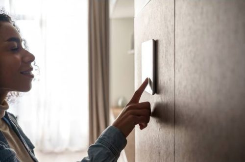 A woman uses a finger to touch the screen of a tablet that hangs on the wall. A window is in the background.