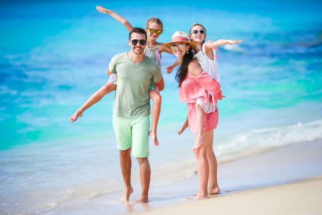 A man and a woman standing on the beach, each carrying a child on their back. They are all wearing sunglasses and smiling.