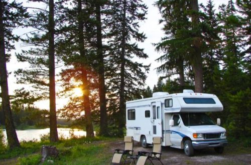 A white RV parked in a heavily forested area near a body of water. There are two camping chairs set up outside the RV.