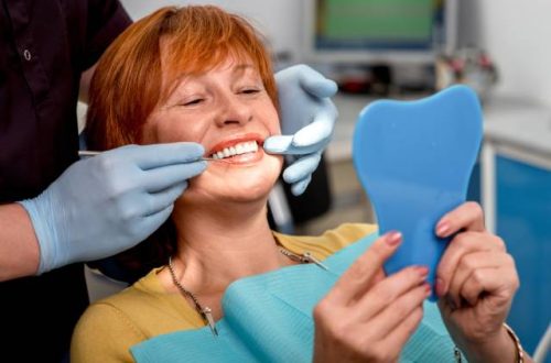 A senior woman is sitting in a dentist chair and holding a small mirror while a person with blue gloves inspects her teeth.
