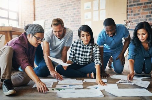 A group of two women and three men are sitting on the floor and organizing paperwork with graphs and charts.