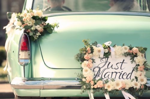 A bride and groom are sitting inside a vintage car. The back of the car has a floral sign that says "Just Married."
