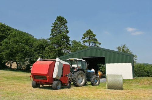 The exterior of a green and white barn with a large tractor and barrel of hay in front of the entrance.
