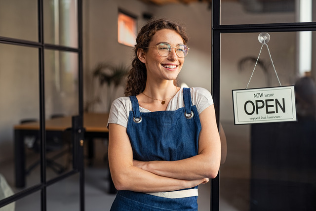 A woman wearing a blue apron stands in the entryway of a building. A sign reads, "Now we are open; support local businesses."