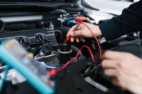 A car mechanic working with the car's hood open. They're performing maintenance on electrical components.
