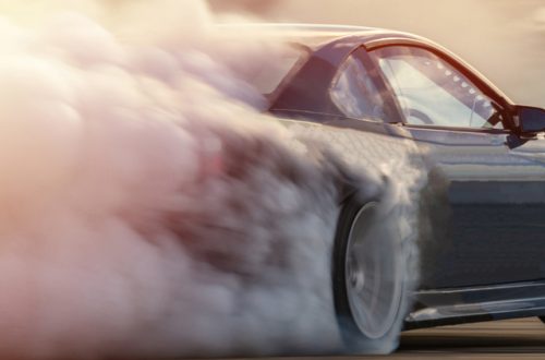 A dark-colored car is creating a lot of smoke by drifting around the corner of a race track. The background is blurry.