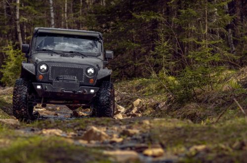 A black off-roading SUV drives on a rocky path that contains water. Tall trees surround the path.