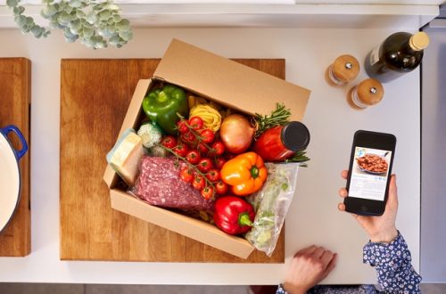 A person holds a smartphone near an open box of recipe ingredients that's part of a meal kit membership.