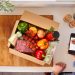 A person holds a smartphone near an open box of recipe ingredients that's part of a meal kit membership.