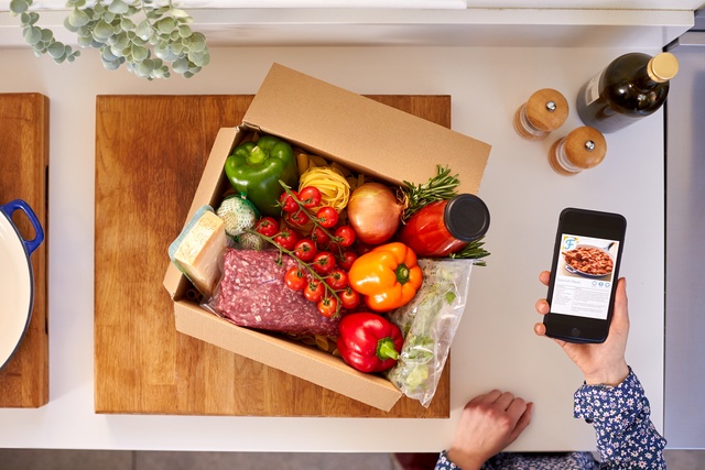A person holds a smartphone near an open box of recipe ingredients that's part of a meal kit membership.