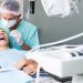 A male dentist in blue scrubs, gloves, and a hair net using tools to treat a female patient's mouth.