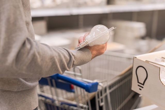 A person wearing a gray sweater holds a packaged light bulb while pushing a shopping cart in a hardware store.