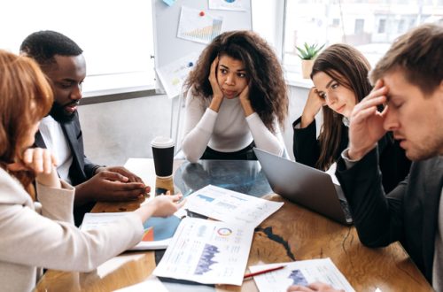 A diverse group of employees sits at a table. Some of them hold their head with a frustrated facial expression.