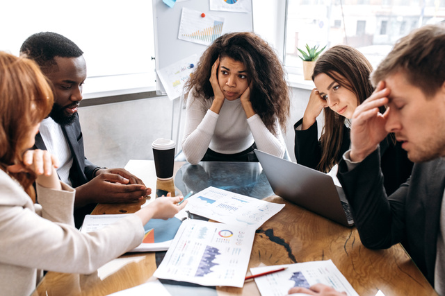 A diverse group of employees sits at a table. Some of them hold their head with a frustrated facial expression.