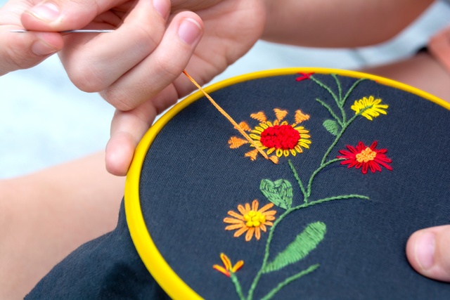 A close-up view shows a hand pulling a needle threaded with yellow thread through a piece of blue fabric with flowers.