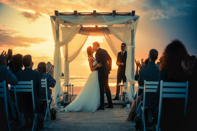 A kissing bride and groom marry on the beach at sunset. Their guests sit on white chairs and watch the ceremony.