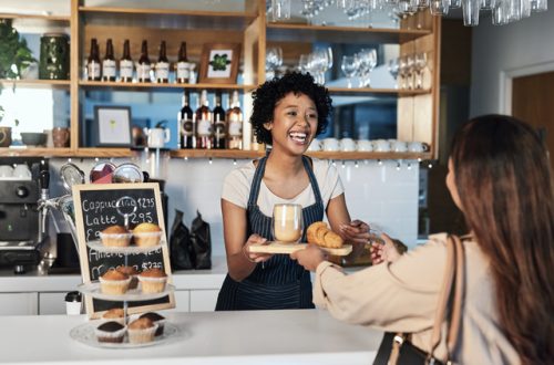 A smiling barista hands a customer her order in a cafe. A latte and croissant sit atop the small wooden tray.