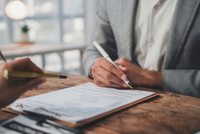 A man in a white shirt and a gray blazer sitting at a wooden table signing paperwork on a clipboard.