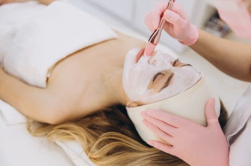 A woman in a towel and headband lies in front of an aesthetician, who paints a white face mask on her face.