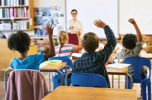 The rear view of young students sitting at desks and raising their hands. A teacher points towards a student.