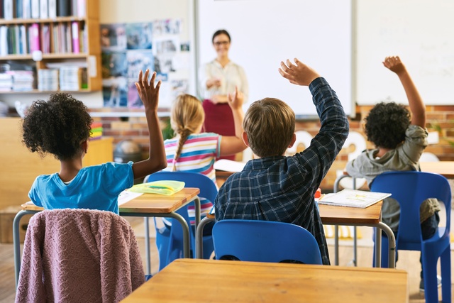 The rear view of young students sitting at desks and raising their hands. A teacher points towards a student.