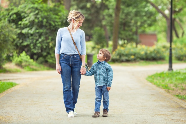 A blonde-haired woman walks hand in hand with a little boy, both strolling together along a paved path outdoors.