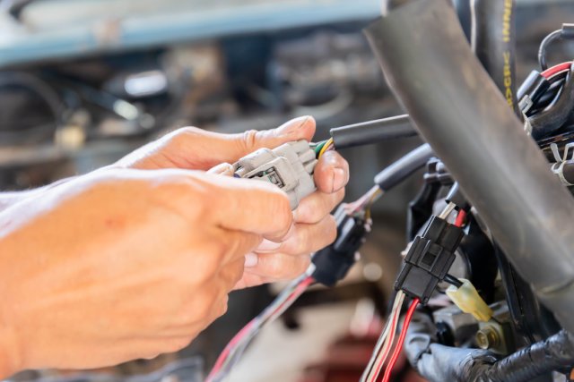 A close-up shows a person's hands holding a gray automotive connector with green and yellow wires coming out of it.