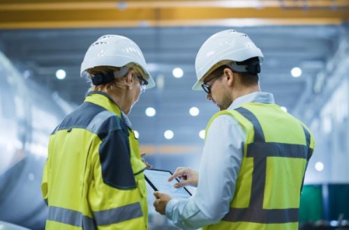 Two engineers wearing white hard hats and high-visibility vests stand next to each other while looking at a tablet.