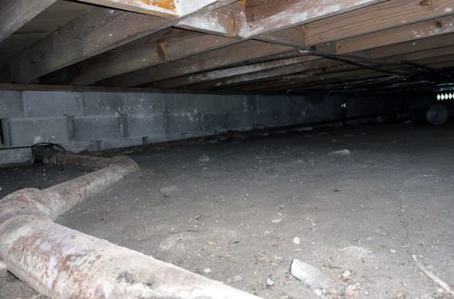 An old, dark crawl space under a home with older, wooden floor joists and dirt on the ground. There is also a long pipe.