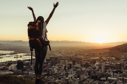 A woman wearing a loaded hiking backpack reaches her arms towards the sky and basks in the view of a cityscape below.