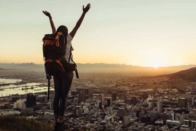 A woman wearing a loaded hiking backpack reaches her arms towards the sky and basks in the view of a cityscape below.