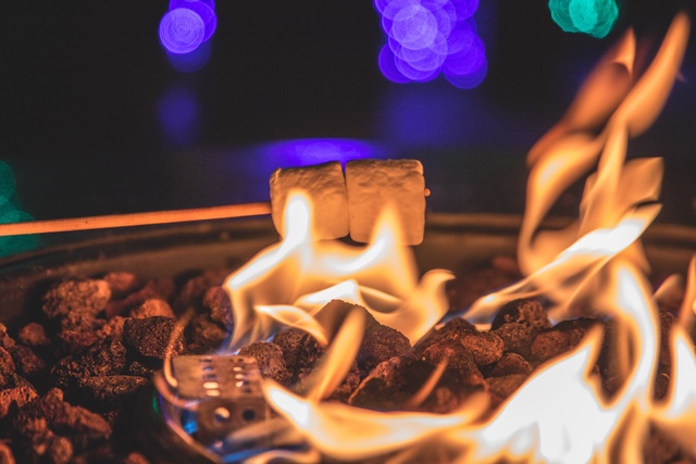 A close-up of a fall bonfire burning in the night. There is a fire stick holding two marshmallows over the flame.