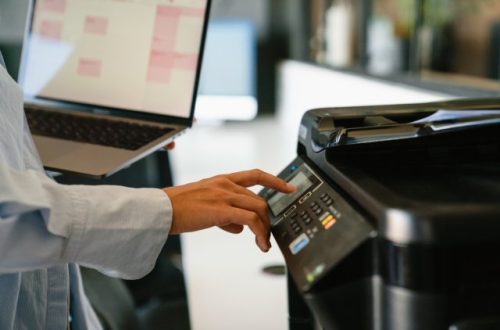 A close-up view shows a person holding a silver laptop in one hand while pushing the button on a printer with the other.