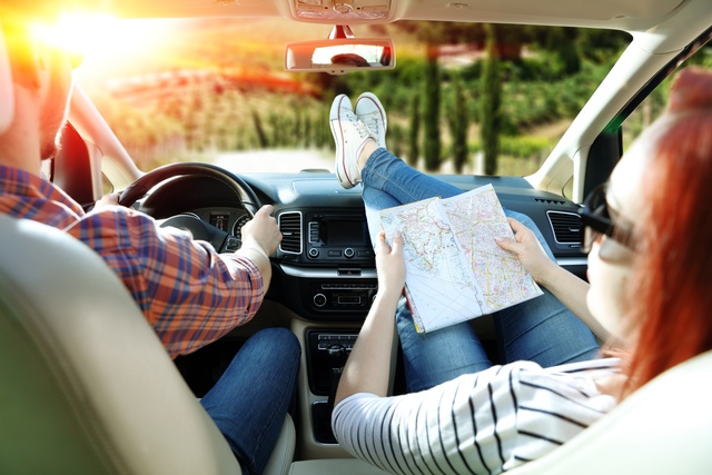 A couple sits together in the front seat of a vehicle as the man drives. The woman has her feet on the dashboard.