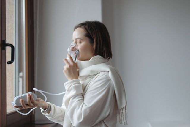 A woman wearing a white shirt and scarf stands in front of a window with her eyes closed, using a portable oxygen machine.