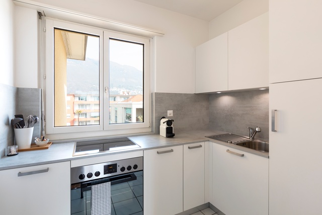 A small, modern white kitchen with a window looking out at the city. There are plenty of cabinets but no pantry.