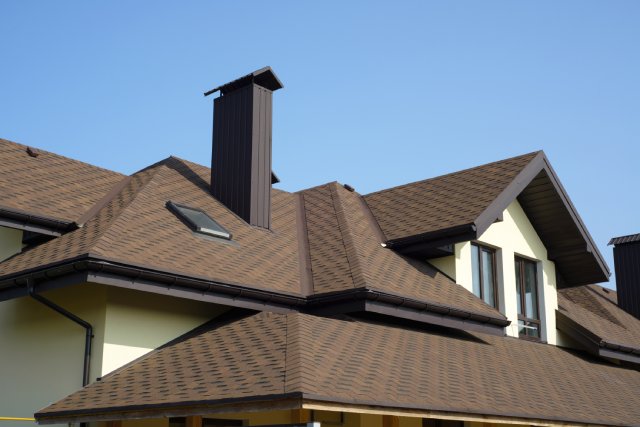 A brand-new, multi-level residential roof with brown shingles, a tall chimney, and a small rectangle skylight.