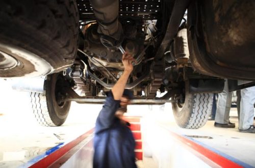 A mechanic wearing a blue jumpsuit stands underneath a lifted vehicle, inspecting the underside with a wrench.